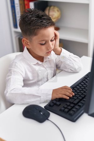 Téléchargez les photos : Adorable élève hispanique garçon utilisant un ordinateur avec une expression fatiguée en classe - en image libre de droit