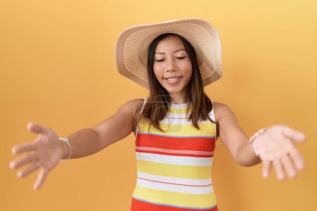Téléchargez les photos : Femme chinoise du Moyen Âge portant un chapeau d'été sur fond jaune regardant la caméra souriant à bras ouverts pour un câlin. expression joyeuse embrassant le bonheur. - en image libre de droit