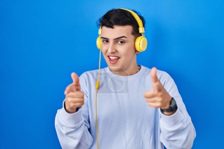 Foto de Non binary person listening to music using headphones pointing fingers to camera with happy and funny face. good energy and vibes. - Imagen libre de derechos