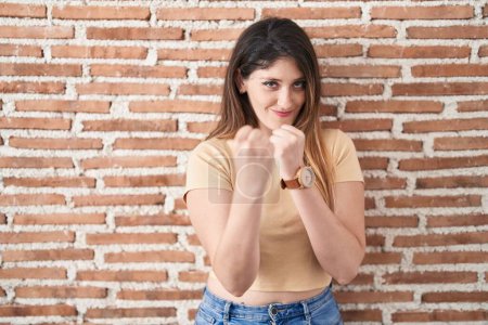 Photo for Young brunette woman standing over bricks wall ready to fight with fist defense gesture, angry and upset face, afraid of problem - Royalty Free Image