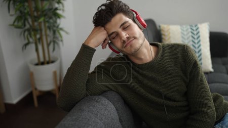 Téléchargez les photos : Jeune homme hispanique écoutant de la musique détendue sur le canapé à la maison - en image libre de droit