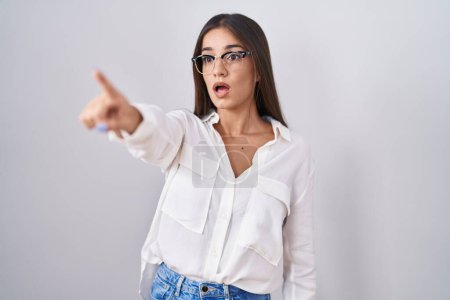Téléchargez les photos : Jeune femme brune portant des lunettes pointant du doigt surpris devant, bouche ouverte expression étonnante, quelque chose sur le devant - en image libre de droit