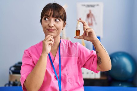Photo for Young brunette woman holding cbd oil at physiotherapy clinic serious face thinking about question with hand on chin, thoughtful about confusing idea - Royalty Free Image