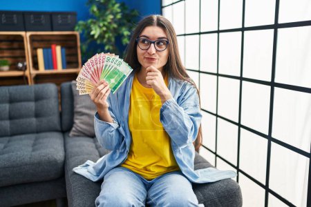 Photo for Young hispanic woman holding norwegian banknotes serious face thinking about question with hand on chin, thoughtful about confusing idea - Royalty Free Image