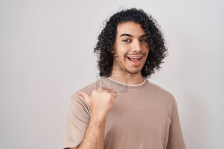 Photo for Hispanic man with curly hair standing over white background smiling with happy face looking and pointing to the side with thumb up. - Royalty Free Image