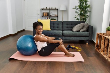 Téléchargez les photos : Femme afro-américaine souriante confiante assise sur un tapis de yoga à la maison - en image libre de droit