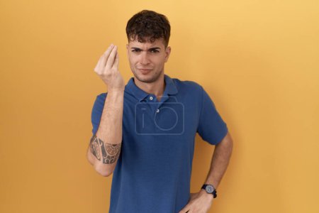 Foto de Young hispanic man standing over yellow background doing italian gesture with hand and fingers confident expression - Imagen libre de derechos