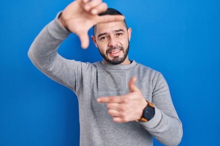 Foto de Hispanic man standing over blue background smiling making frame with hands and fingers with happy face. creativity and photography concept. - Imagen libre de derechos