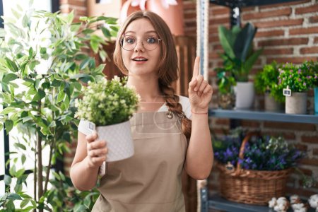 Photo for Young caucasian woman working at florist shop holding plant smiling with an idea or question pointing finger with happy face, number one - Royalty Free Image