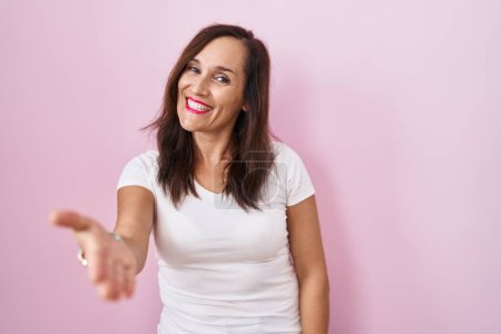 Foto de Middle age brunette woman standing over pink background smiling friendly offering handshake as greeting and welcoming. successful business. - Imagen libre de derechos