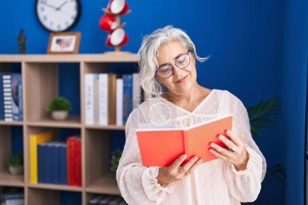 Foto de Mujer de pelo gris de mediana edad leyendo libro de pie en casa - Imagen libre de derechos