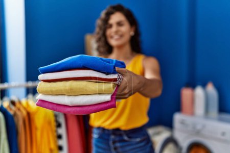 Foto de Young latin woman smiling confident holding folded clothes at laundry room - Imagen libre de derechos