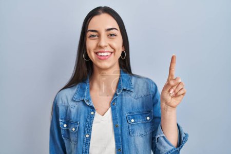 Téléchargez les photos : Femme hispanique debout sur fond bleu montrant et pointant vers le haut avec le doigt numéro un tout en souriant confiant et heureux. - en image libre de droit