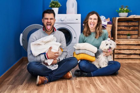 Téléchargez les photos : Jeune couple hispanique faisant la lessive assis sur le sol langue collante heureux avec une expression drôle. - en image libre de droit