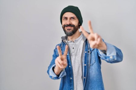 Téléchargez les photos : Jeune homme hispanique avec des tatouages portant un bonnet en laine souriant en regardant la caméra montrant des doigts faisant signe de victoire. numéro deux. - en image libre de droit