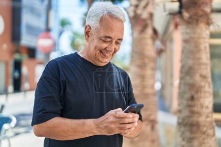 Téléchargez les photos : Moyen âge homme aux cheveux gris souriant confiant en utilisant smartphone à la rue - en image libre de droit