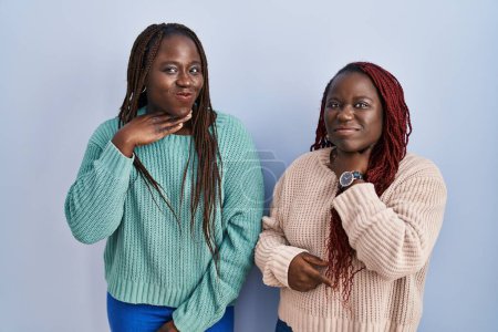 Foto de Dos mujeres africanas de pie sobre fondo azul cortando la garganta con la mano como cuchillo, amenazan la agresión con violencia furiosa - Imagen libre de derechos