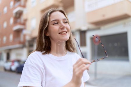 Foto de Joven mujer caucásica sonriendo confiado sosteniendo gafas en la calle - Imagen libre de derechos