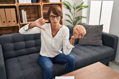Photo for Brunette woman working at therapy office holding brain with angry face, negative sign showing dislike with thumbs down, rejection concept - Royalty Free Image