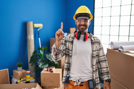 Foto de Young hispanic man with beard working at home renovation showing and pointing up with finger number one while smiling confident and happy. - Imagen libre de derechos