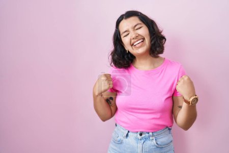 Foto de Mujer hispana joven de pie sobre fondo rosa muy feliz y emocionada haciendo gesto ganador con los brazos levantados, sonriendo y gritando por el éxito. concepto de celebración. - Imagen libre de derechos