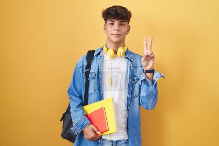 Téléchargez les photos : Adolescent hispanique portant un sac à dos d'étudiant et tenant des livres souriant avec un clin d'œil heureux à la caméra faisant signe de victoire avec les doigts. numéro deux. - en image libre de droit