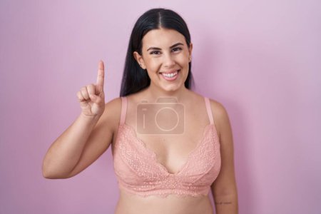 Téléchargez les photos : Jeune femme hispanique portant un soutien-gorge rose montrant et pointant vers le haut avec le doigt numéro un tout en souriant confiant et heureux. - en image libre de droit