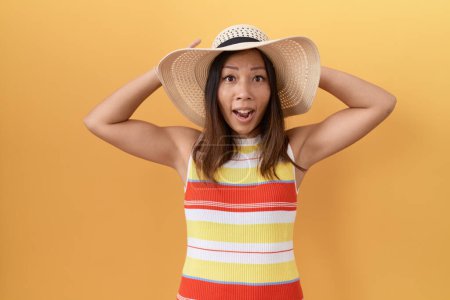 Foto de Middle age chinese woman wearing summer hat over yellow background crazy and scared with hands on head, afraid and surprised of shock with open mouth - Imagen libre de derechos