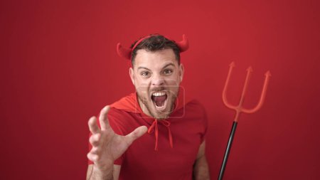Téléchargez les photos : Jeune homme caucasien criant portant costume diable sur fond rouge isolé - en image libre de droit