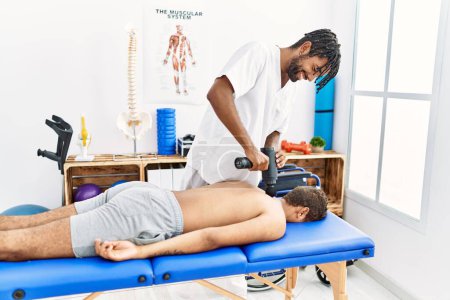 Foto de Dos hombres fisioterapeuta y paciente masajeando la espalda usando pistola de percusión en la clínica - Imagen libre de derechos