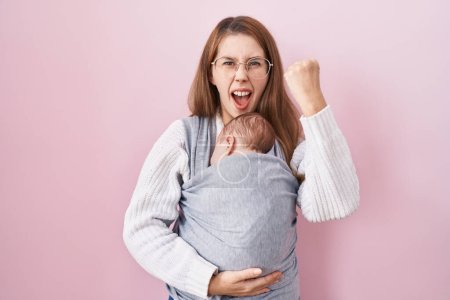 Foto de Joven mujer caucásica sosteniendo y cargando al bebé en un cabestrillo molesto y frustrado gritando con ira, gritando loca de ira y mano levantada - Imagen libre de derechos