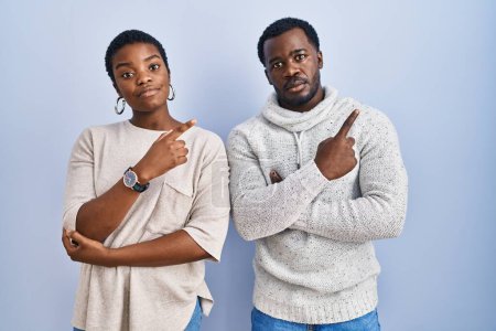 Téléchargez les photos : Jeune couple afro-américain debout sur fond bleu pointant ensemble avec le doigt de la main sur le côté montrant la publicité, visage sérieux et calme - en image libre de droit