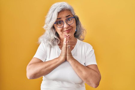 Photo for Middle age woman with grey hair standing over yellow background praying with hands together asking for forgiveness smiling confident. - Royalty Free Image