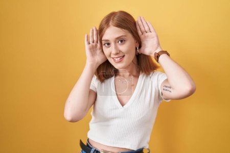 Téléchargez les photos : Jeune femme rousse debout sur fond jaune essayant d'entendre les deux mains sur le geste de l'oreille, curieuse de commérages. problème auditif, sourd - en image libre de droit