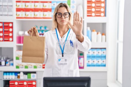 Photo for Young blonde woman working at pharmacy drugstore holding paper bag with open hand doing stop sign with serious and confident expression, defense gesture - Royalty Free Image