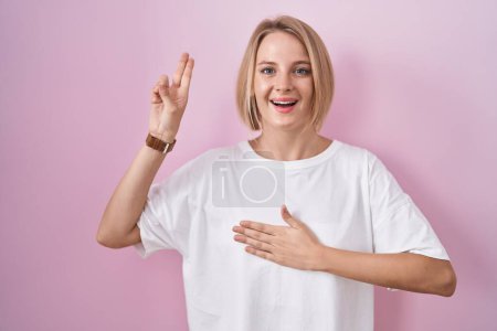 Foto de Mujer caucásica joven de pie sobre fondo rosa sonriendo jurando con la mano en el pecho y los dedos hacia arriba, haciendo una promesa de lealtad juramento - Imagen libre de derechos