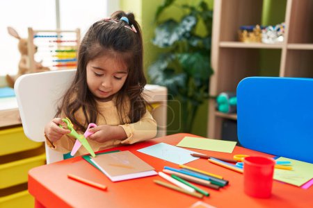 Photo for Adorable hispanic girl student sitting on table cutting paper at kindergarten - Royalty Free Image