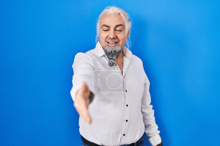 Photo for Middle age man with grey hair standing over blue background smiling friendly offering handshake as greeting and welcoming. successful business. - Royalty Free Image