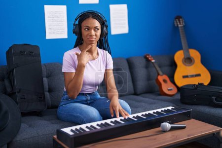 Foto de Mujer afroamericana con trenzas tocando el teclado de piano en el estudio de música cara seria pensando en la pregunta con la mano en la barbilla, pensativo acerca de la idea confusa - Imagen libre de derechos