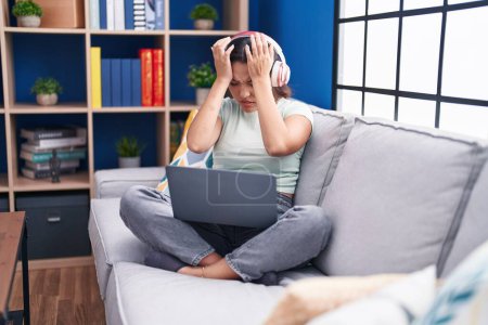 Photo for Hispanic young woman using laptop at home wearing headphones suffering from headache desperate and stressed because pain and migraine. hands on head. - Royalty Free Image