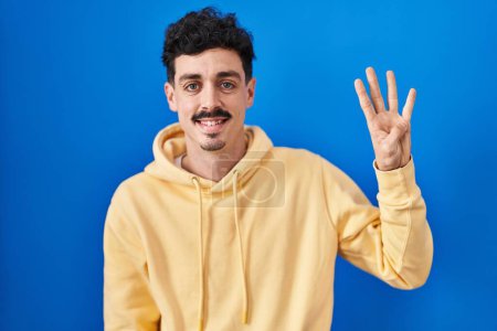 Foto de Hombre hispano de pie sobre fondo azul mostrando y señalando hacia arriba con los dedos número cuatro mientras sonríe confiado y feliz. - Imagen libre de derechos