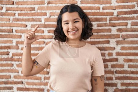 Téléchargez les photos : Jeune femme hispanique debout sur le mur de briques geste souriant et confiant avec la main faisant signe de petite taille avec les doigts regardant et la caméra. concept de mesure. - en image libre de droit