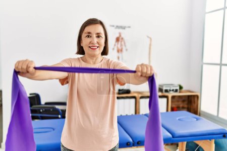 Téléchargez les photos : Femme hispanique d'âge moyen faisant de l'exercice avec des bandes élastiques à la clinique de physiothérapie - en image libre de droit