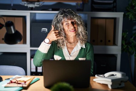 Photo for Middle age woman working at night using computer laptop smiling doing phone gesture with hand and fingers like talking on the telephone. communicating concepts. - Royalty Free Image