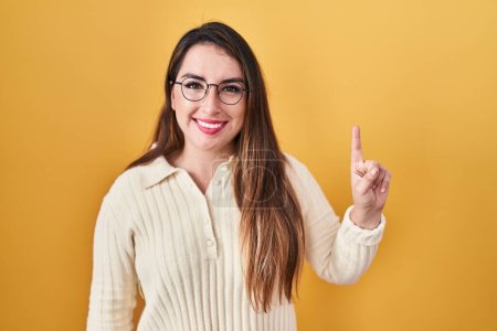 Photo for Young hispanic woman standing over yellow background showing and pointing up with finger number one while smiling confident and happy. - Royalty Free Image