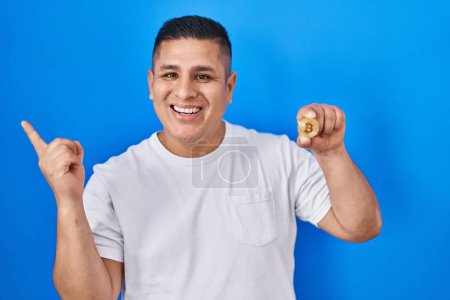 Foto de Joven hispano sosteniendo bitcoin moneda virtual sonriendo feliz señalando con la mano y el dedo a un lado - Imagen libre de derechos