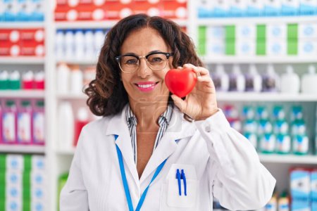 Téléchargez les photos : Middle age woman pharmacist smiling confident holding heart at pharmacy - en image libre de droit