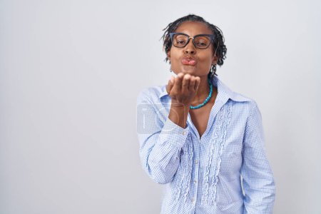 Téléchargez les photos : Femme africaine avec dreadlocks debout sur fond blanc portant des lunettes regardant la caméra souffler un baiser avec la main sur l'air étant belle et sexy. expression d'amour. - en image libre de droit