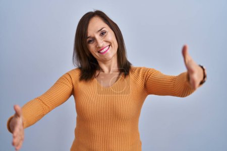 Téléchargez les photos : Middle age brunette woman standing wearing orange sweater looking at the camera smiling with open arms for hug. cheerful expression embracing happiness. - en image libre de droit