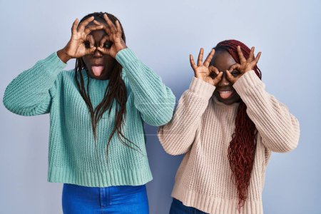 Foto de Dos mujeres africanas paradas sobre un fondo azul haciendo un buen gesto como binoculares sacando la lengua, ojos mirando a través de los dedos. expresión loca. - Imagen libre de derechos